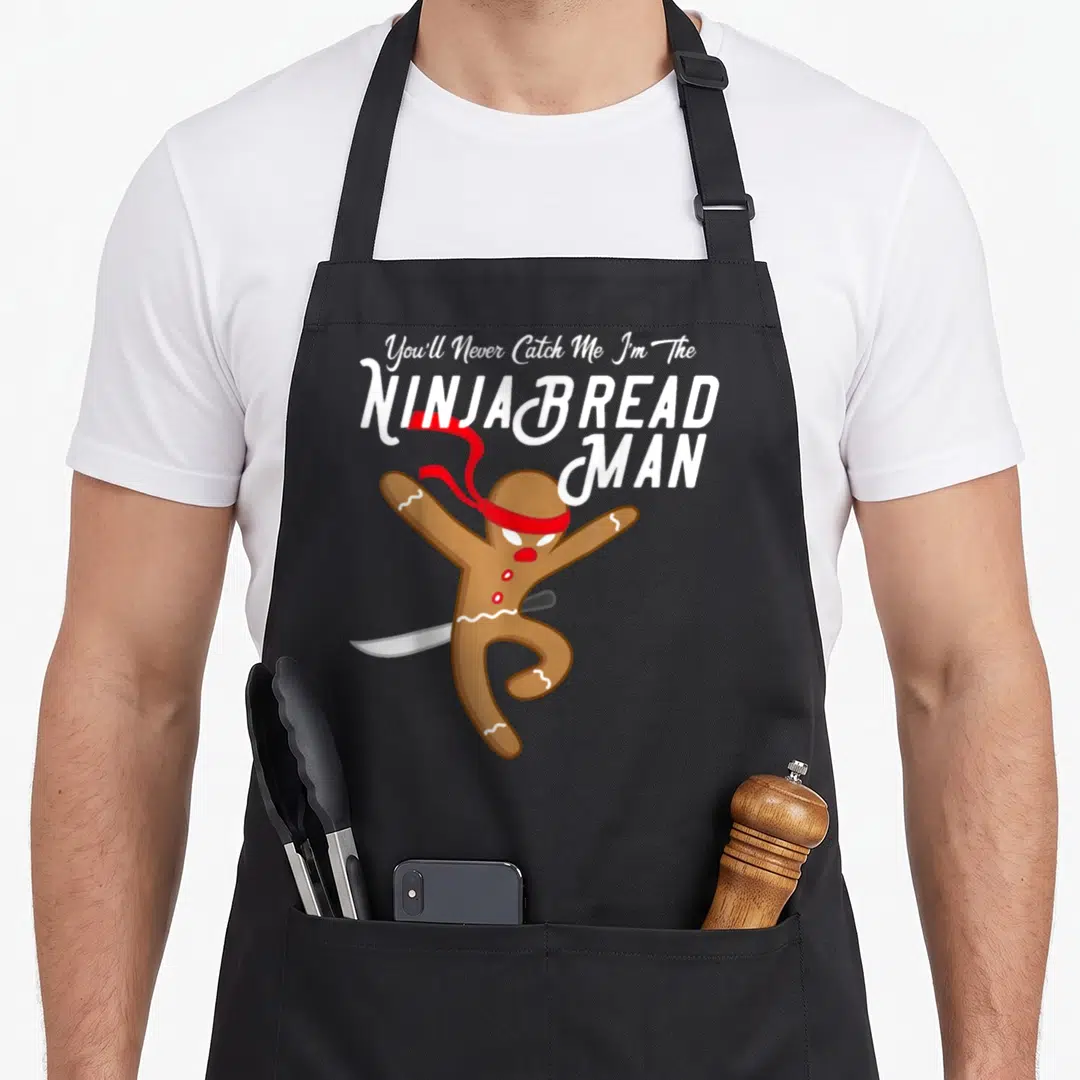 Catch christmas apron - Ninja gingerbread man holding sword with red headband and stacked white text on grey background — shown on a black cotton canvas apron against a white background