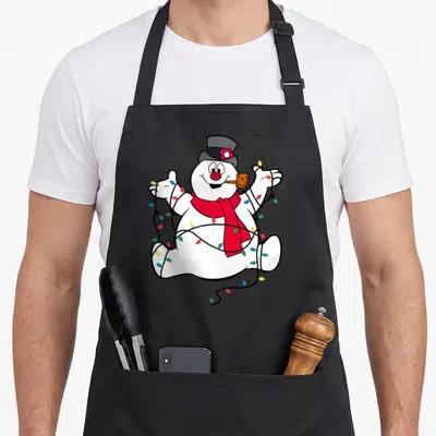 Portrait christmas snowman apron - Cartoon snowman sitting with red scarf, orange pipe, wrapped in multicolored — shown on a black cotton canvas apron against a white background