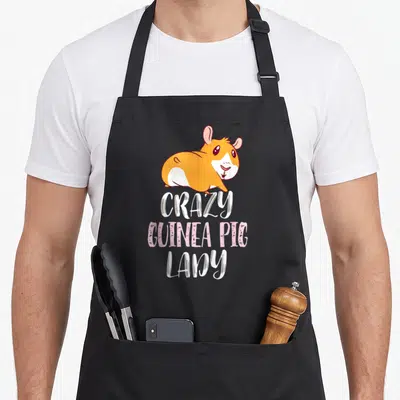 Cool pig crazy guinea lady apron - Cartoon orange and white guinea pig above stacked CRAZY GUINEA PIG LADY text in pink — shown on a black cotton canvas apron against a white background