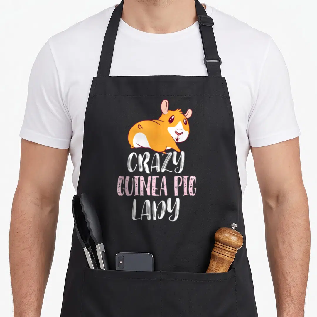 Cool pig crazy guinea lady apron - Cartoon orange and white guinea pig above stacked CRAZY GUINEA PIG LADY text in pink — shown on a black cotton canvas apron against a white background