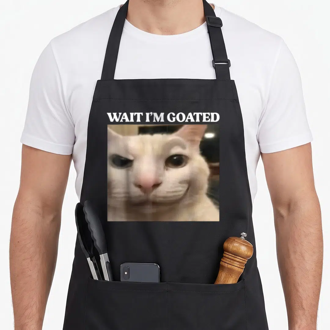 Goated cat apron - White text WAIT I'M GOATED above a square photo of a smirking beige and white cat face — shown on a black cotton canvas apron against a white background