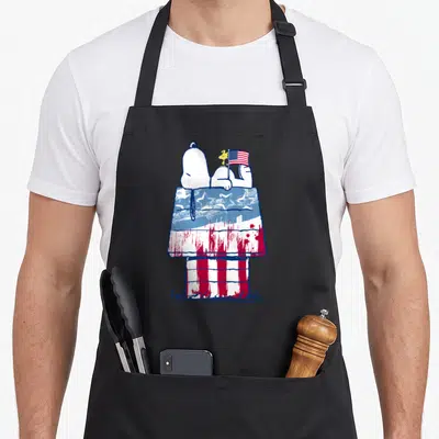 Cute wood apron - Cartoon dog on red white and blue flag doghouse with small bird holding flag — shown on a black cotton canvas apron against a white background