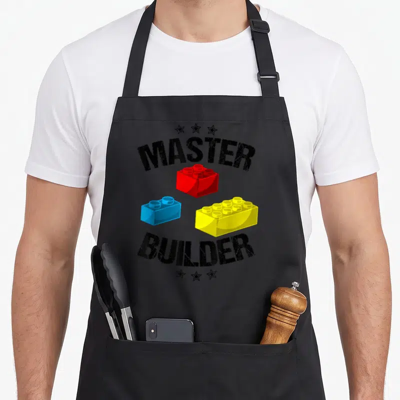 Master building apron - Arched MASTER BUILDER text above red, blue, and yellow building blocks with stars — shown on a black cotton canvas apron against a white background