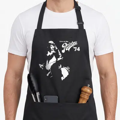 Queen rainbow apron - White silhouette of a man playing guitar next to stacked text reading Live at the Rainbow '74 — shown on a black cotton canvas apron against a white background