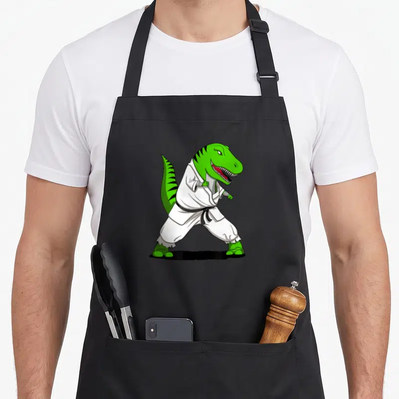 Karate dinosaur apron - Green cartoon T-Rex in white gi and black belt standing in karate stance — shown on a black cotton canvas apron against a white background
