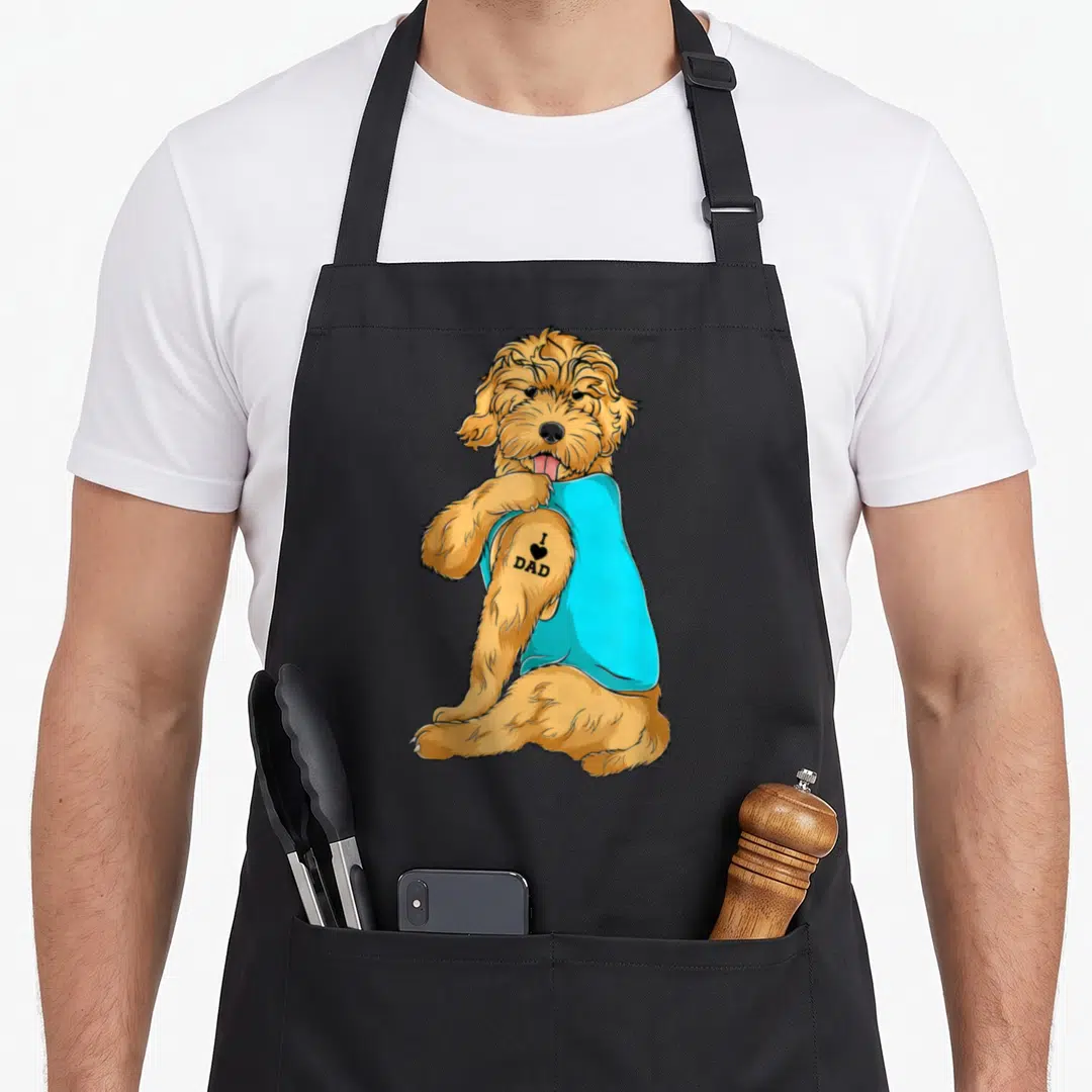 Golden tattoo aprons - Cartoon golden dog in cyan shirt with I heart DAD tattoo on arm — shown on a black cotton canvas apron against a white background