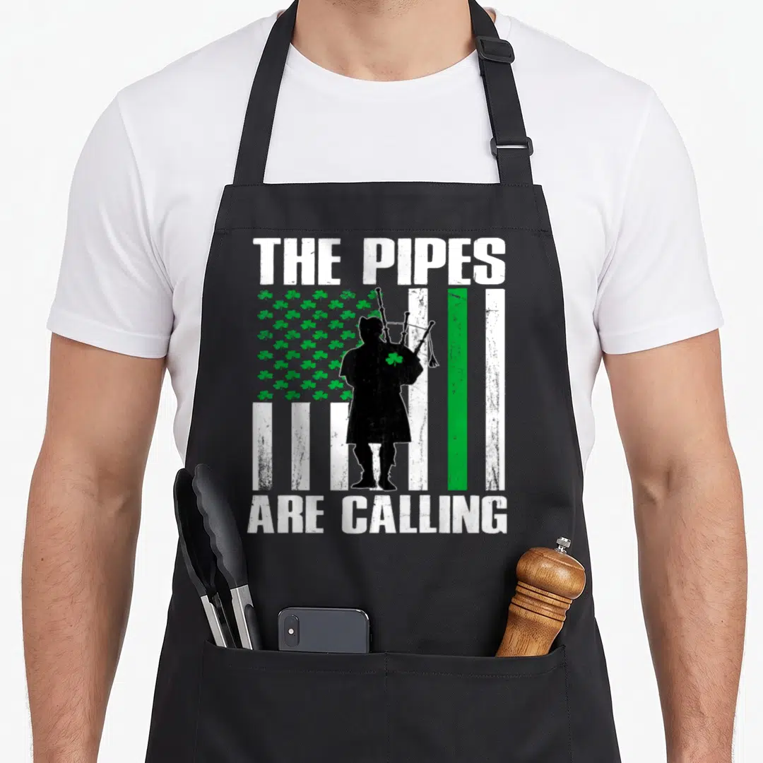 Pipe st patrick day aprons - Distressed bagpiper silhouette over shamrock flag with white text reading The Pipes Are — shown on a black cotton canvas apron against a white background