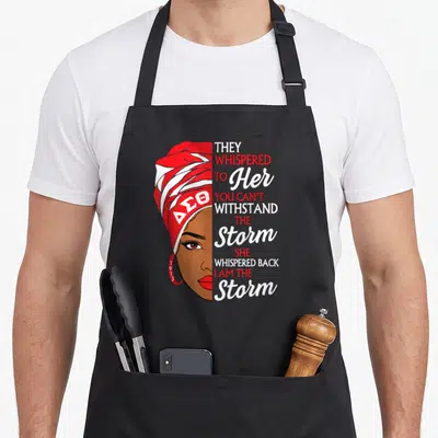 Theta sorority apron - Woman in red headwrap with DSO letters and stacked text reading They Whispered To Her You Can't — shown on a black cotton canvas apron against a white background
