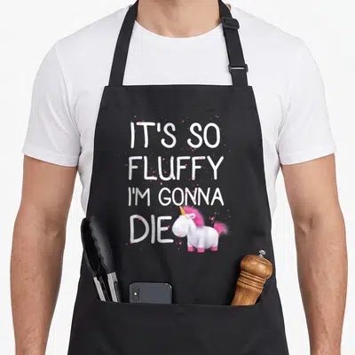 Despicable unicorn apron - White text IT'S SO FLUFFY I'M GONNA DIE above pink and white cartoon unicorn with pink — shown on a black cotton canvas apron against a white background