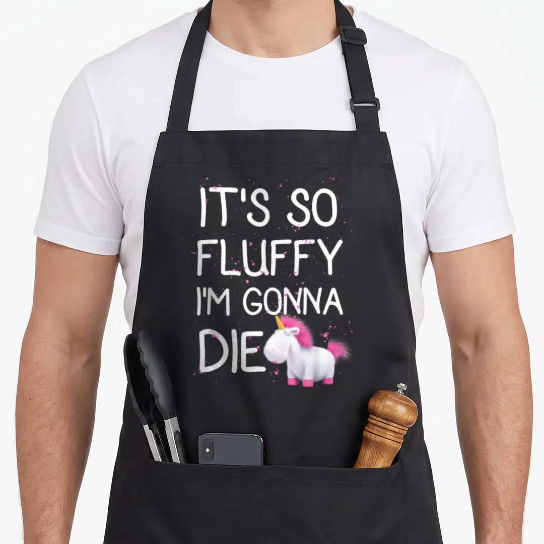 Despicable unicorn apron - White text IT'S SO FLUFFY I'M GONNA DIE above pink and white cartoon unicorn with pink — shown on a black cotton canvas apron against a white background