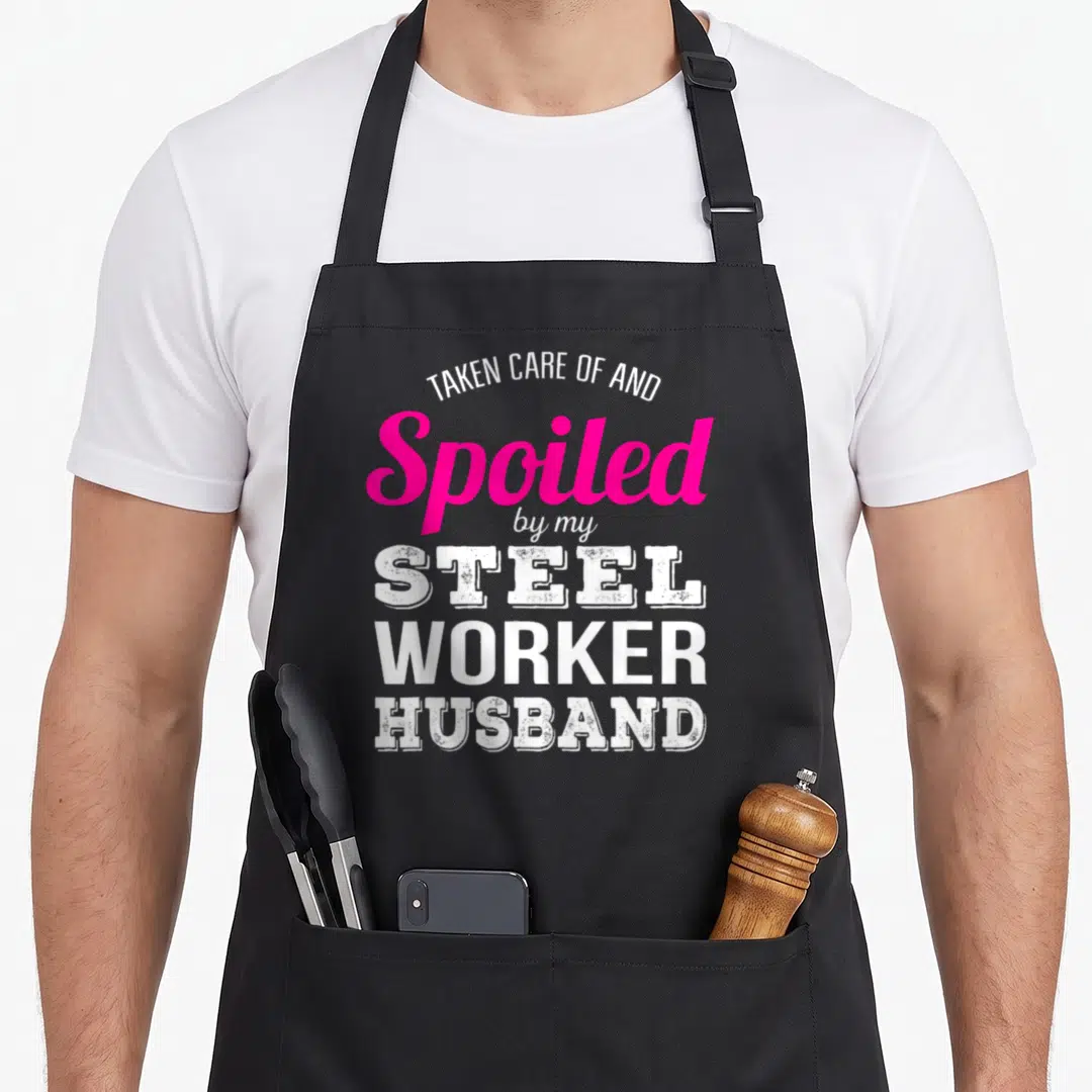 Steel wife apron - Stacked white and pink text reading Taken Care Of And Spoiled By My Steel Worker Husband — shown on a black cotton canvas apron against a white background