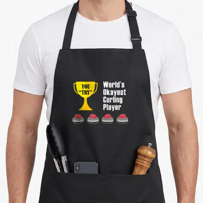 Okayest curling apron - Yellow trophy with YOU TRY text and four red-handled curling stones on grey background — shown on a black cotton canvas apron against a white background