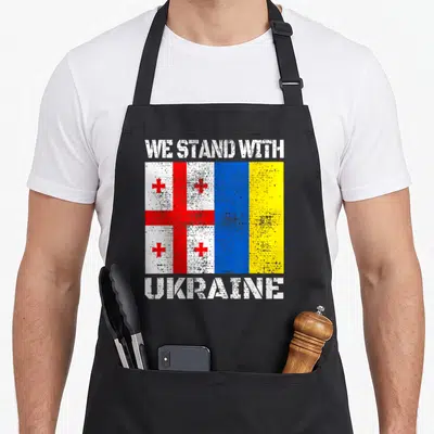 Georgian ukrainian aprons - We Stand With Ukraine text flanking side-by-side Georgian and Ukrainian flags in distressed — shown on a black cotton canvas apron against a white background