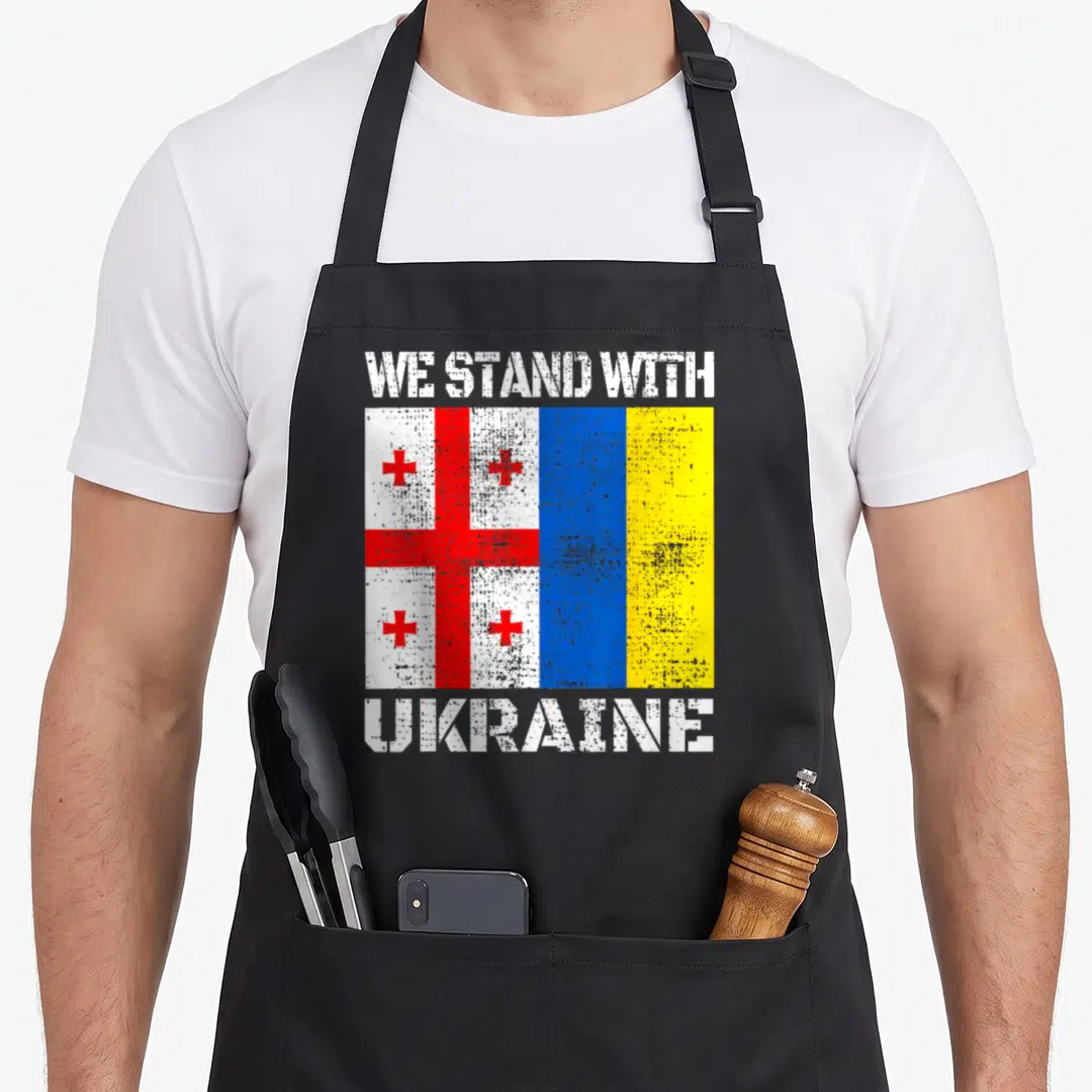 Georgian ukrainian aprons - We Stand With Ukraine text flanking side-by-side Georgian and Ukrainian flags in distressed — shown on a black cotton canvas apron against a white background