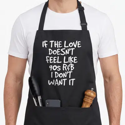 Feel 90s r aprons - White stacked text reading If the Love Doesn't Feel Like 90s R&B I Don't Want It — shown on a black cotton canvas apron against a white background
