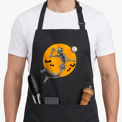 Volleyball halloween apron - Cartoon skeleton jumping for a volleyball inside an orange circle with four black bats — shown on a black cotton canvas apron against a white background