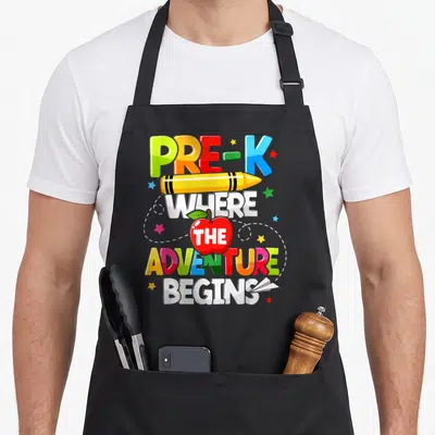 Pre adventure teacher apron - Colorful PRE-K text with yellow crayon and red apple containing THE surrounded by stars — shown on a black cotton canvas apron against a white background