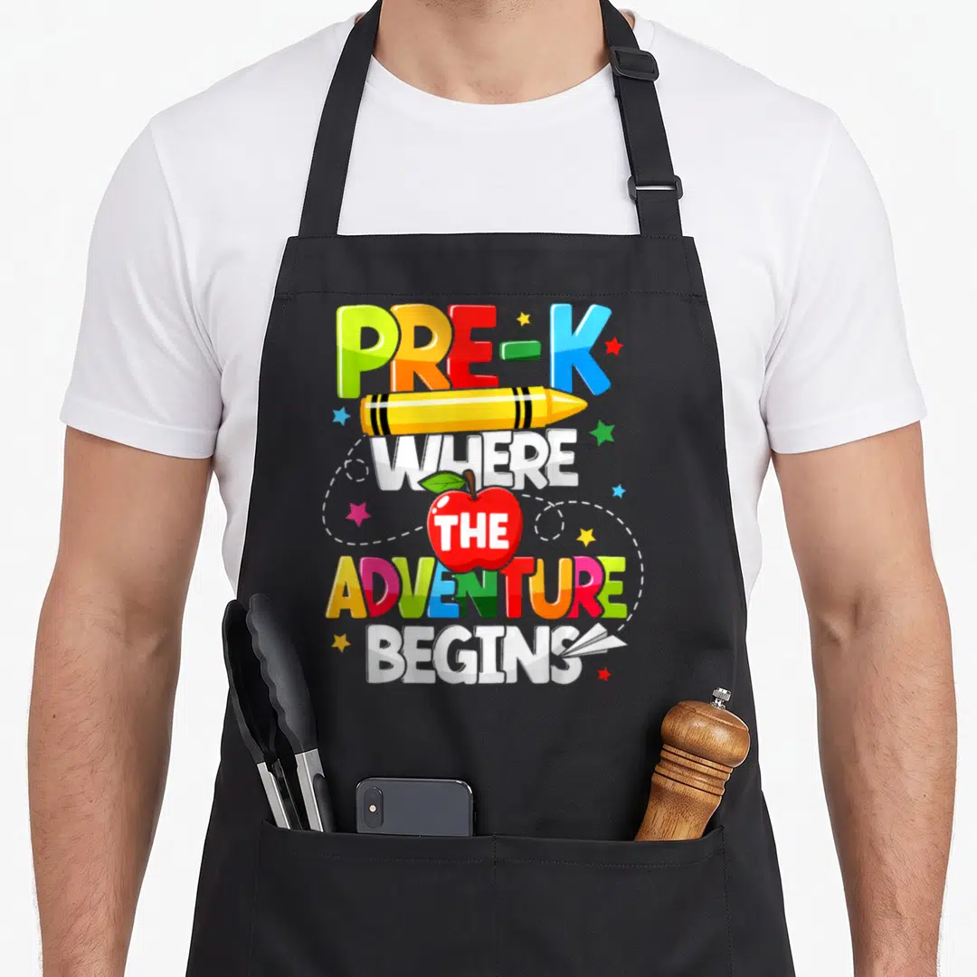 Pre adventure teacher apron - Colorful PRE-K text with yellow crayon and red apple containing THE surrounded by stars — shown on a black cotton canvas apron against a white background