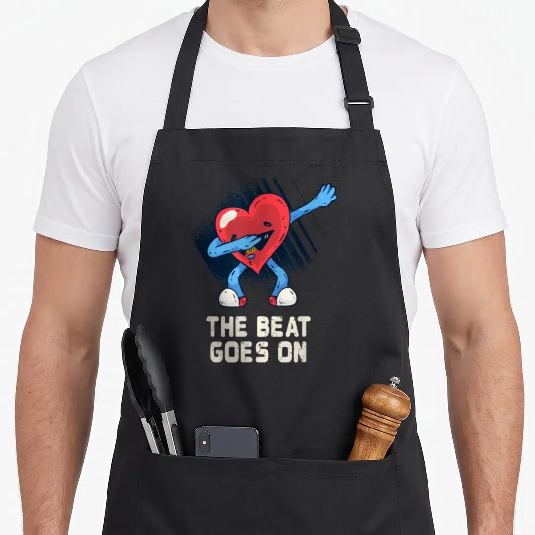Surgery survivor open apron - Dancing red heart character with blue limbs and beige text reading THE BEAT GOES — shown on a black cotton canvas apron against a white background