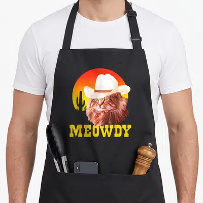 Meowdy cook's country aprons - Brown cat wearing white cowboy hat in front of yellow red gradient sun with cactus — shown on a black cotton canvas apron against a white background