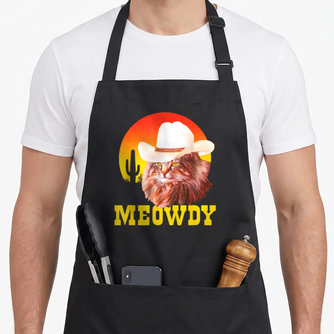 Meowdy cook's country aprons - Brown cat wearing white cowboy hat in front of yellow red gradient sun with cactus — shown on a black cotton canvas apron against a white background