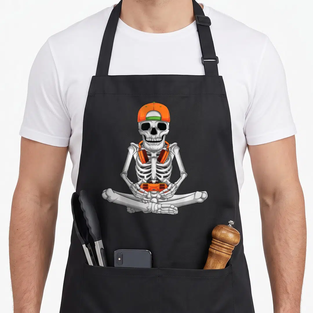 Video halloween apron - Cartoon skeleton sitting cross-legged holding orange controller wearing orange cap — shown on a black cotton canvas apron against a white background