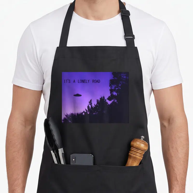 Lonely road apron - Black UFO and trees on purple sky with IT'S A LONELY ROAD text — shown on a black cotton canvas apron against a white background