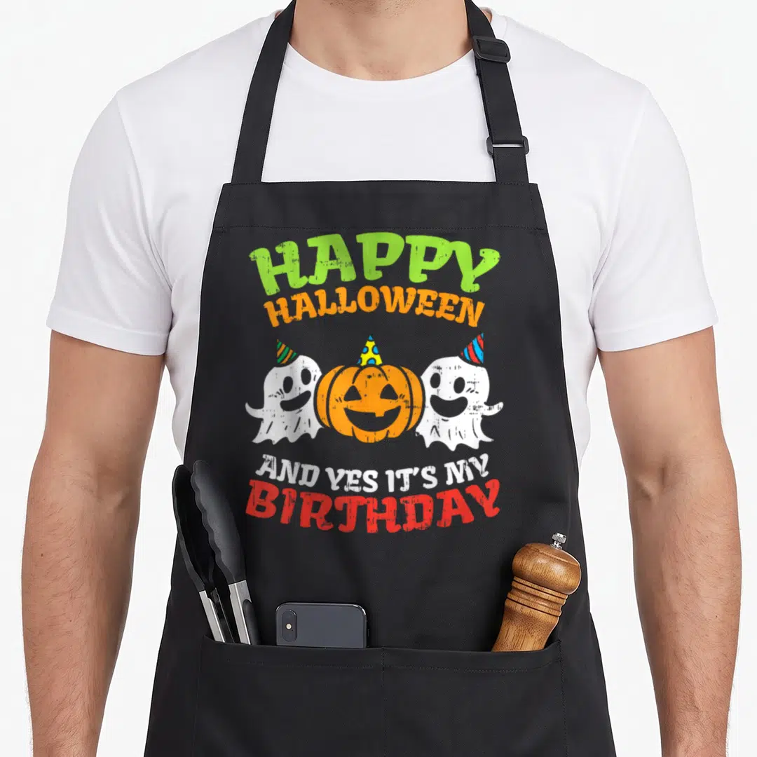 Funny birthday funny halloween apron - Cartoon pumpkin between two white ghosts wearing party hats with green orange — shown on a black cotton canvas apron against a white background