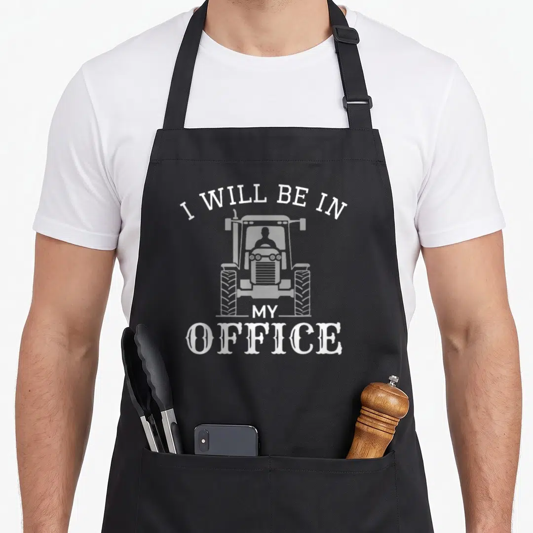 Office farmer apron - White tractor graphic with text I Will Be In My Office arched above and stacked below — shown on a black cotton canvas apron against a white background