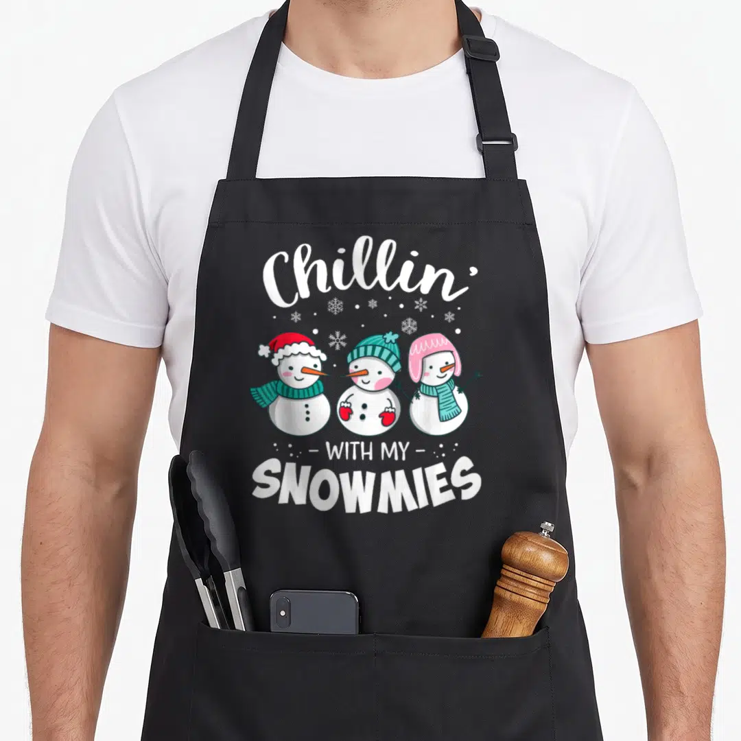 Snowmie snowman apron - Three cartoon snowmen in teal red and pink below Chillin with My Snowmies text — shown on a black cotton canvas apron against a white background