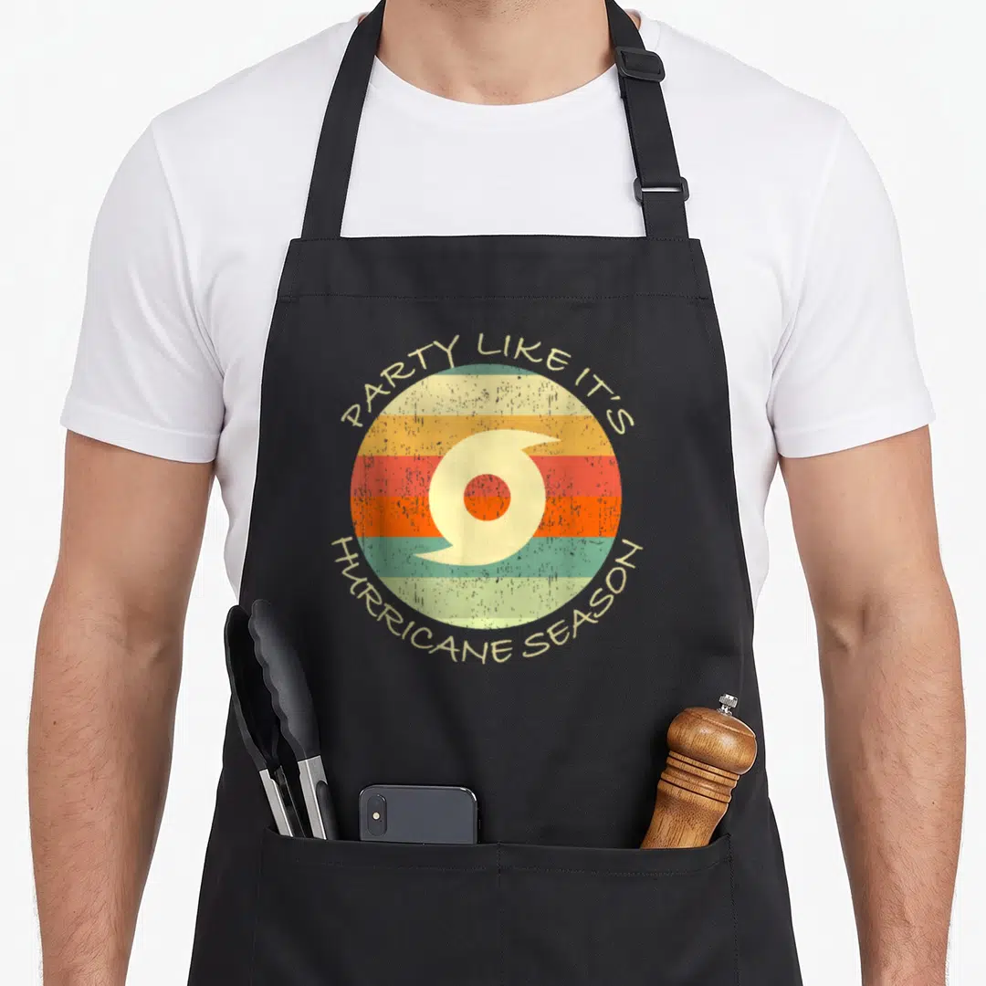 Season hurricane apron - Retro striped circle with hurricane symbol and arching text PARTY LIKE IT'S HURRICANE SEASON — shown on a black cotton canvas apron against a white background