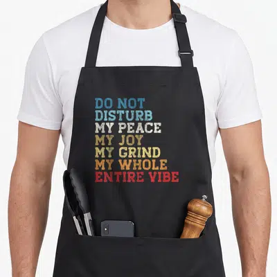 Joy grind apron - Stacked DO NOT DISTURB text in distressed blue white gold and red typography — shown on a black cotton canvas apron against a white background