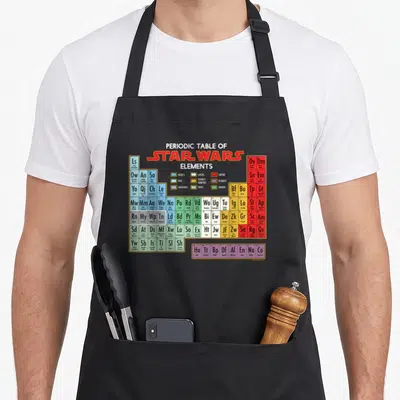 Periodic table apron - Star Wars Elements grid with characters ships and planets in colored cells — shown on a black cotton canvas apron against a white background