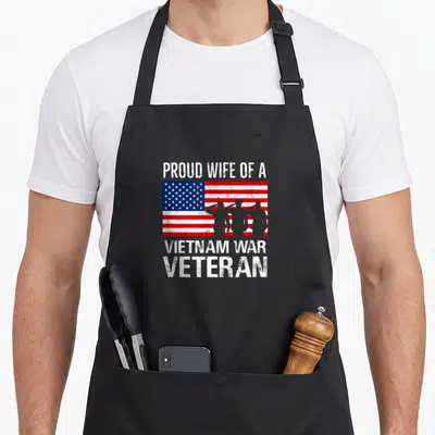 Wife best husband apron - Distressed USA flag with three saluting soldier silhouettes and white text reading PROUD WIFE — shown on a black cotton canvas apron against a white background
