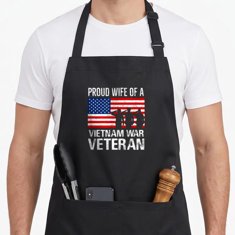 Wife best husband apron - Distressed USA flag with three saluting soldier silhouettes and white text reading PROUD WIFE — shown on a black cotton canvas apron against a white background