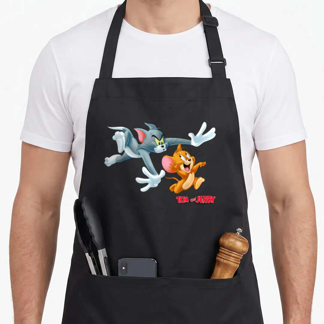 Chase novelty apron - Tom the cat lunges at Jerry the mouse with TOM and JERRY text below — shown on a black cotton canvas apron against a white background