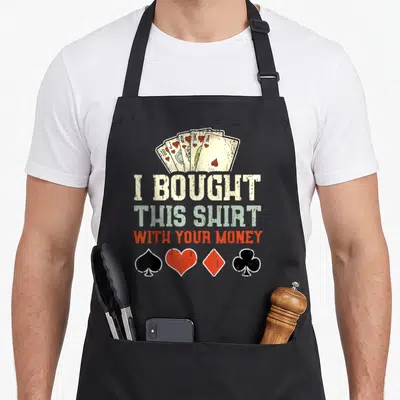 Bought money apron - Fan of hearts above stacked text I BOUGHT THIS SHIRT WITH YOUR MONEY and four suit symbols — shown on a black cotton canvas apron against a white background