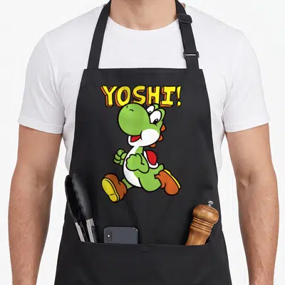 Intro jump apron - Arched YOSHI! text above a walking green cartoon dinosaur with white belly — shown on a black cotton canvas apron against a white background