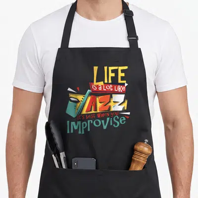 Sax band humor apron - Stacked colorful text reading Life Is a Lot Like Jazz Improvise with geometric shapes in yellow — shown on a black cotton canvas apron against a white background