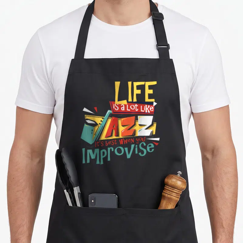 Sax band humor apron - Stacked colorful text reading Life Is a Lot Like Jazz Improvise with geometric shapes in yellow — shown on a black cotton canvas apron against a white background