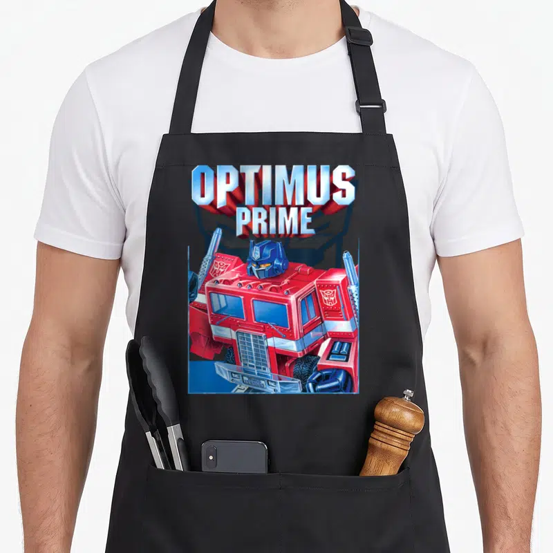 Funny portrait transformer apron - OPTIMUS PRIME 3D text above red blue silver cartoon truck cab facing forward — shown on a black cotton canvas apron against a white background