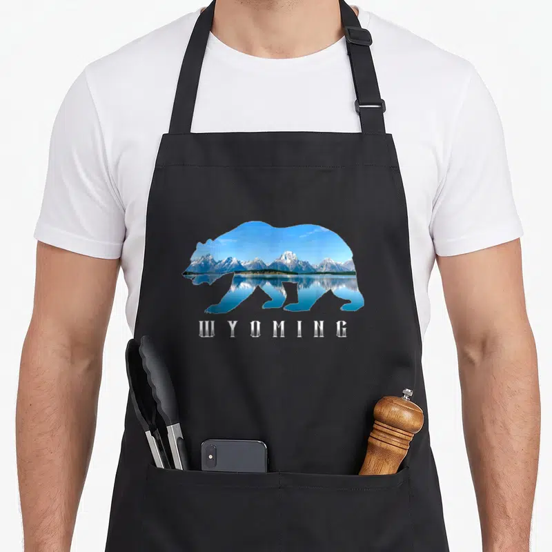 Wyoming bear apron - Bear silhouette filled with blue mountain lake photo and white WYOMING text — shown on a black cotton canvas apron against a white background