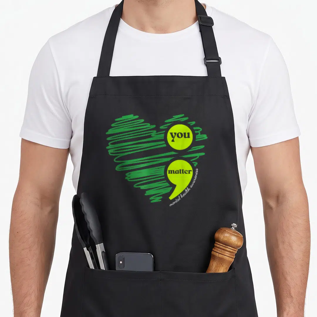 Matter limes apron - Scribbled green heart with lime circle and comma shapes containing text you matter — shown on a black cotton canvas apron against a white background