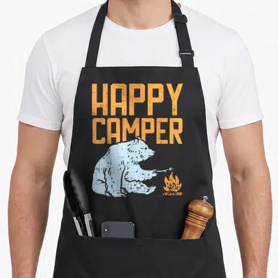Hiking happy camper apron - Orange HAPPY CAMPER text above light blue bear roasting marshmallow over campfire — shown on a black cotton canvas apron against a white background