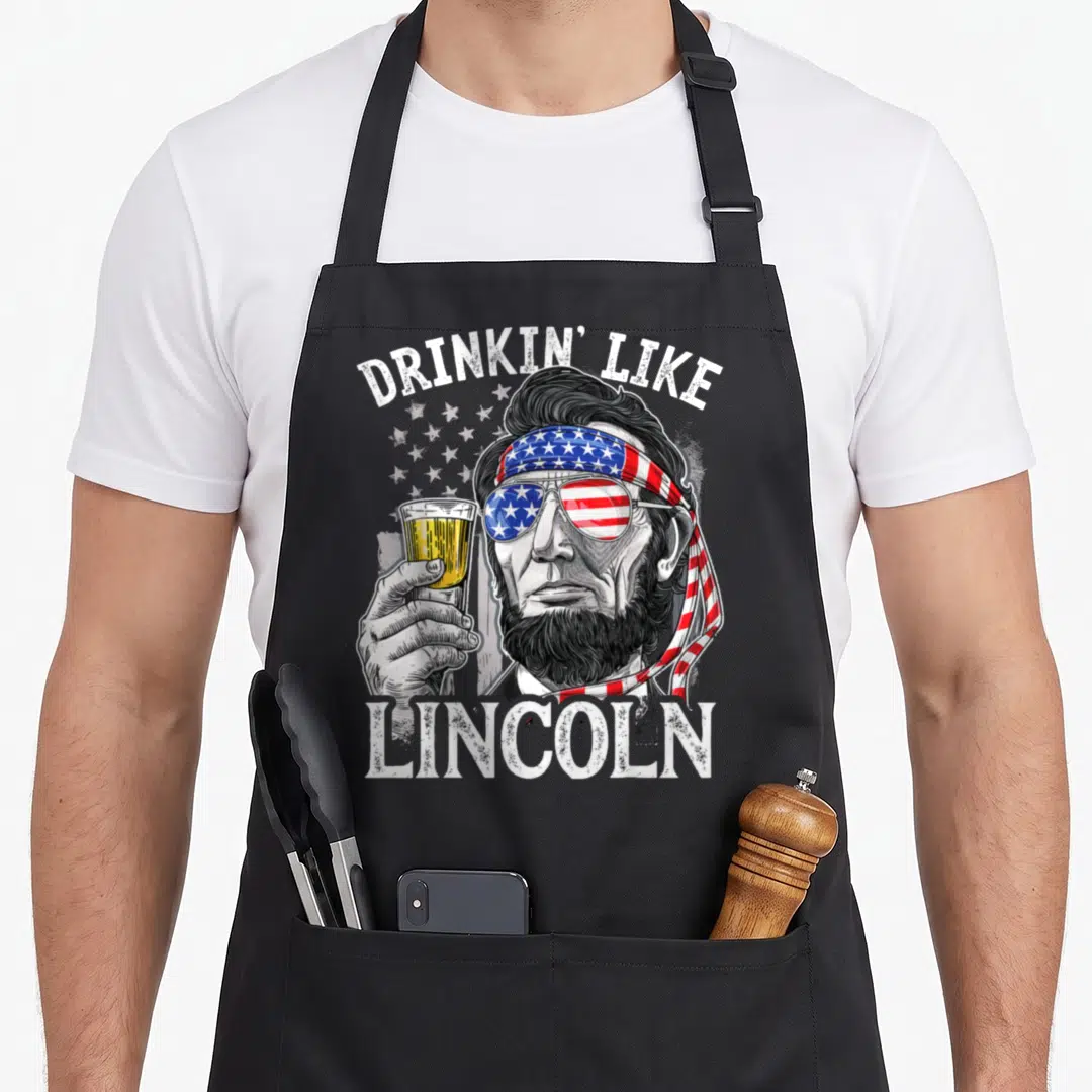 Drinking 4th of july apron - Distressed graphic of Lincoln in flag sunglasses holding beer with arched text — shown on a black cotton canvas apron against a white background