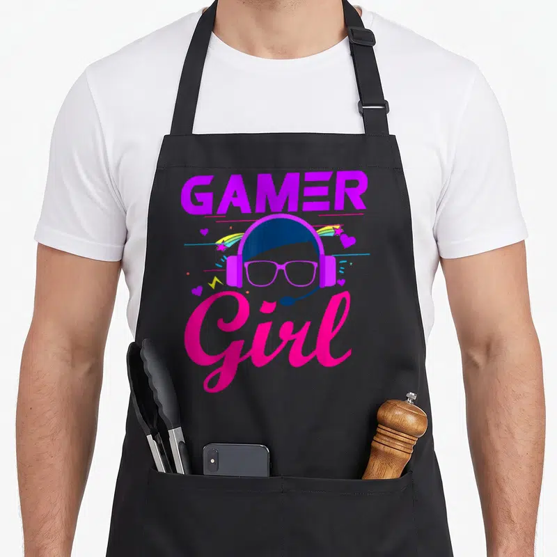 Family video game apron - Gamer Girl text with cartoon avatar wearing headset glasses surrounded by rainbow stars — shown on a black cotton canvas apron against a white background