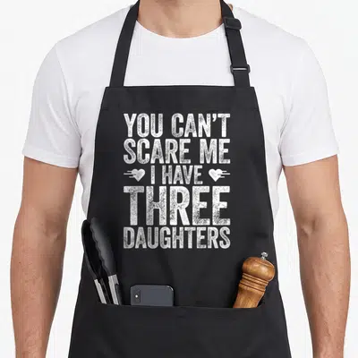 Three mom daughter aprons - White distressed text reading YOU CAN'T SCARE ME I HAVE THREE DAUGHTERS flanked by two — shown on a black cotton canvas apron against a white background