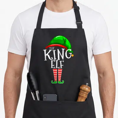 King christmas elf apron - Green elf hat white THE KING ELF text red striped legs green shoes cartoon — shown on a black cotton canvas apron against a white background