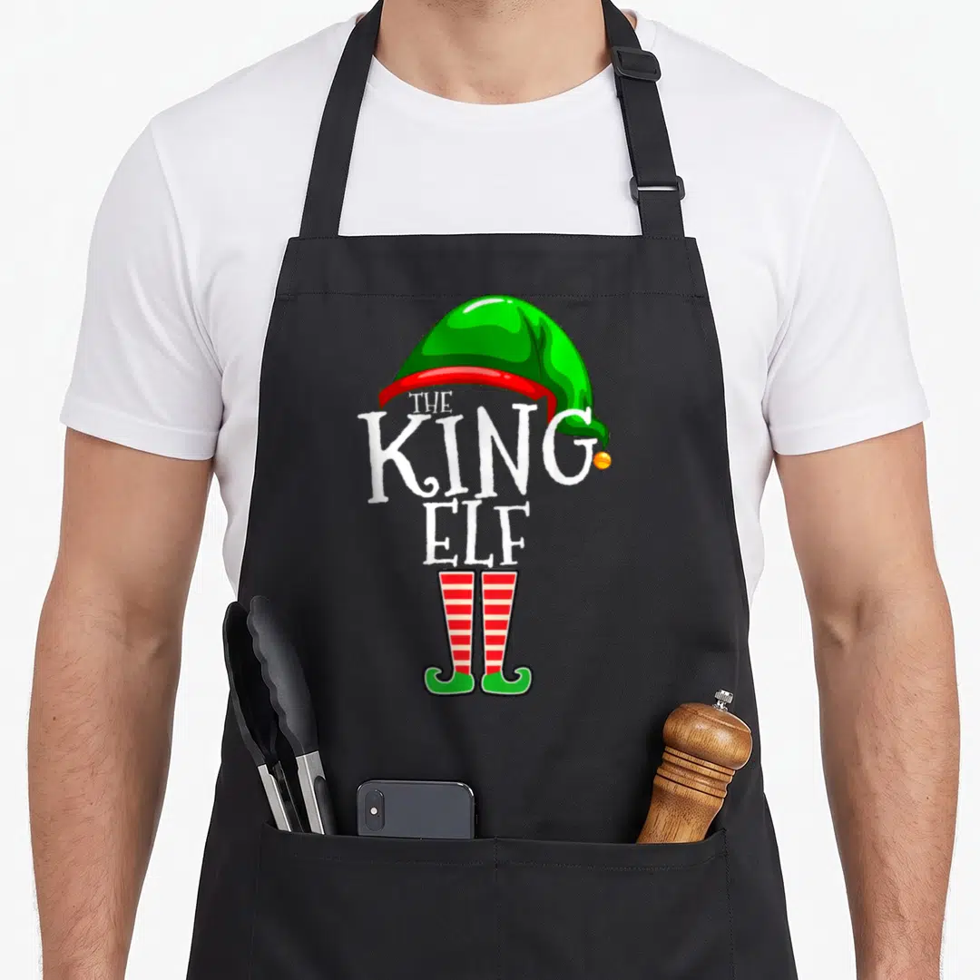 King christmas elf apron - Green elf hat white THE KING ELF text red striped legs green shoes cartoon — shown on a black cotton canvas apron against a white background