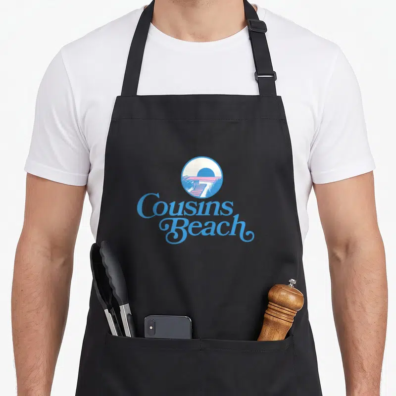 Cousin pretty apron - Retro circular beach scene with blue script reading Cousins Beach — shown on a black cotton canvas apron against a white background